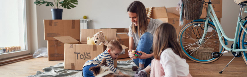 young mom playing with her children and a dog on the wooden floor in new apartment with unpacked boxes around them.